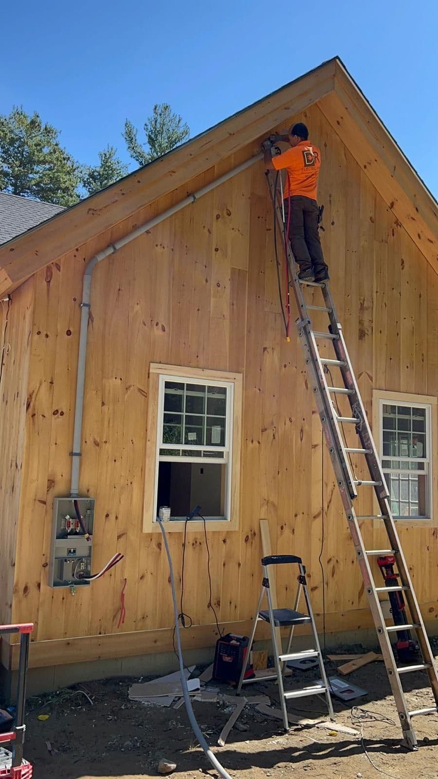 Worker on ladder installing electrical equipment on modern wooden house exterior.