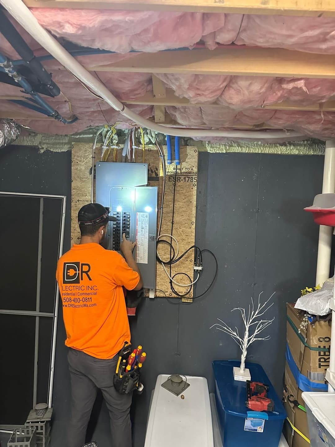 Electrician working on a circuit breaker box in a residential basement setting.