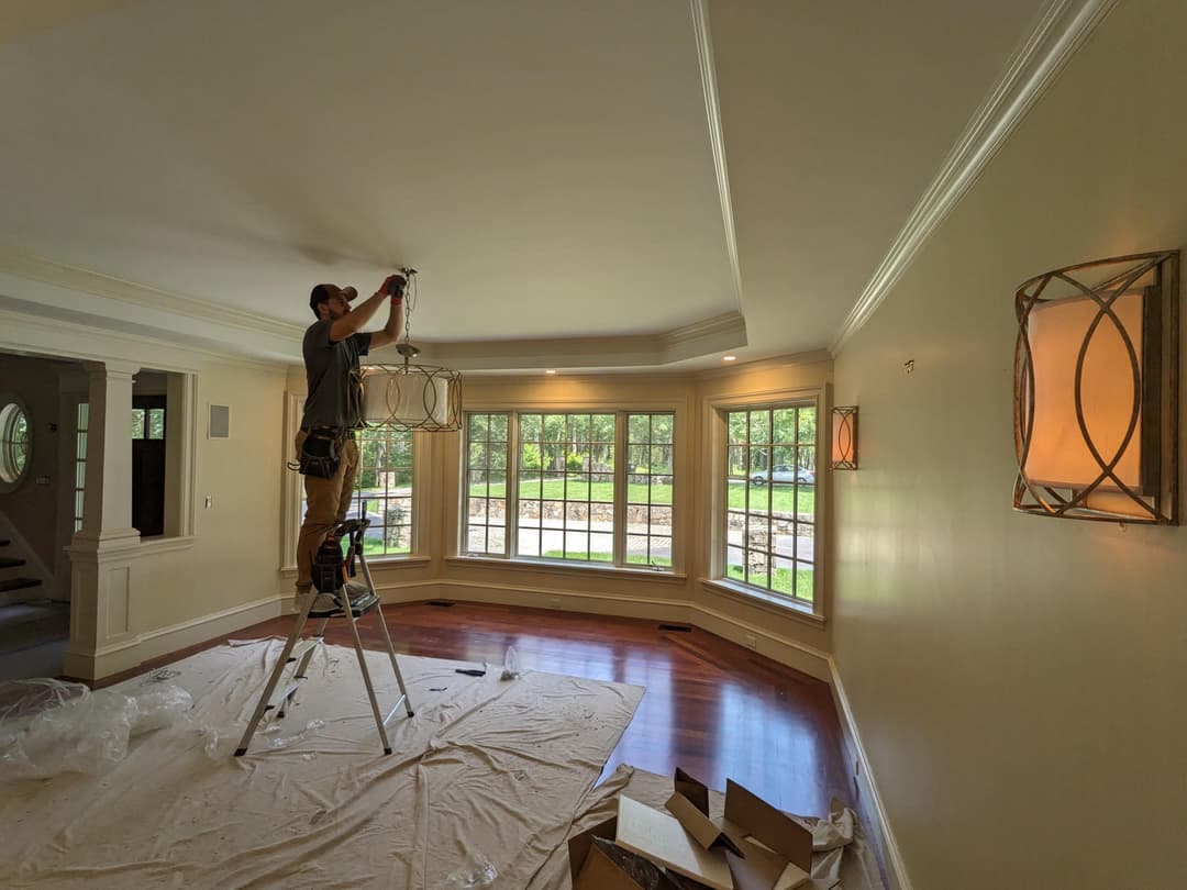 Electrician installing a chandelier in a bright living room with large windows.
